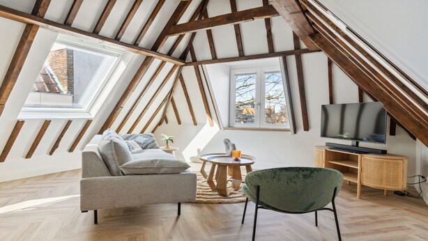 Ein gemütliches Schlafzimmer im Loft-Stil mit freiliegenden Holzbalken, einer Couch, einem Sessel, einem Fernseher und einem Fenster mit Blick auf die Stadt.