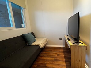Una stanza piccola con un divano dai colori scuri, un televisore su un mobile in legno e una finestra con persiane.