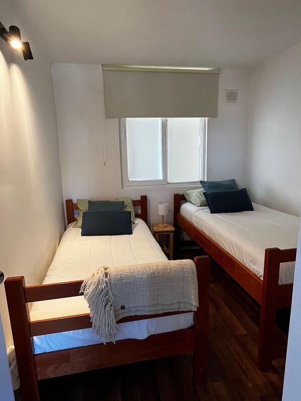 Un dormitorio con dos literas de madera, una ventana con persianas, una mesita de noche pequeña y una lámpara fijada a la pared.