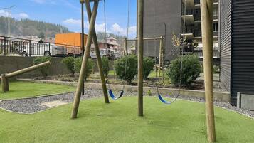 A playground with swings and a climbing structure, surrounded by a well-maintained grassy area and a building with balconies.