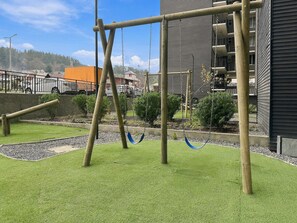 A playground with swings and a climbing structure, surrounded by a well-maintained grassy area and a building with balconies. - Oceana Suites Alto Panorámico (Villarrica)