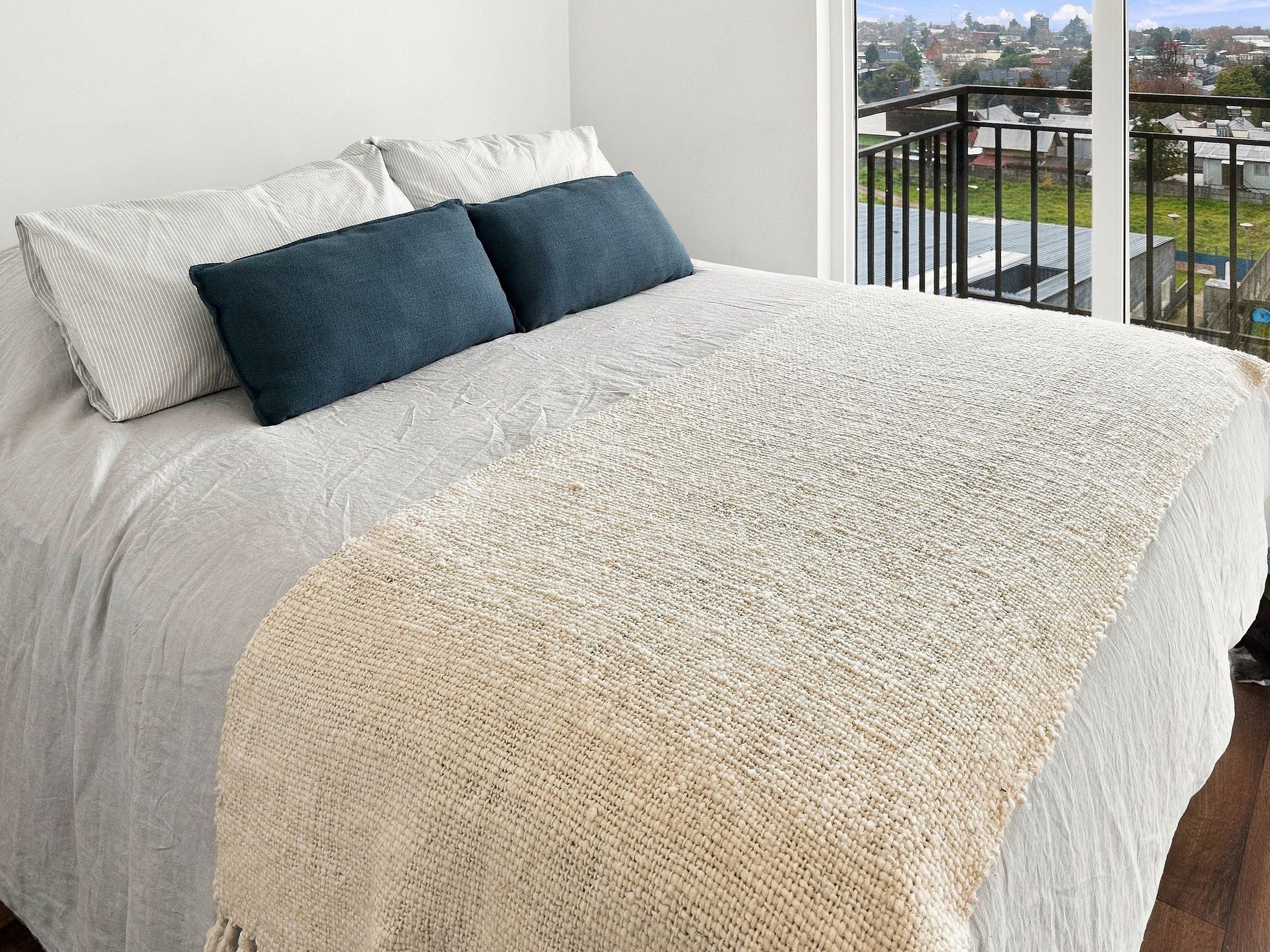 Una cama con una colcha blanca y beige, dos almohadas y vista a un paisaje urbano a través de la barandilla de un balcón.