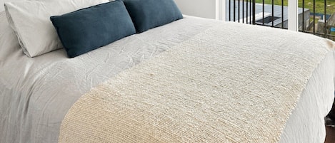 Una cama con una colcha blanca y beige, dos almohadas y vista a un paisaje urbano a través de la barandilla de un balcón.