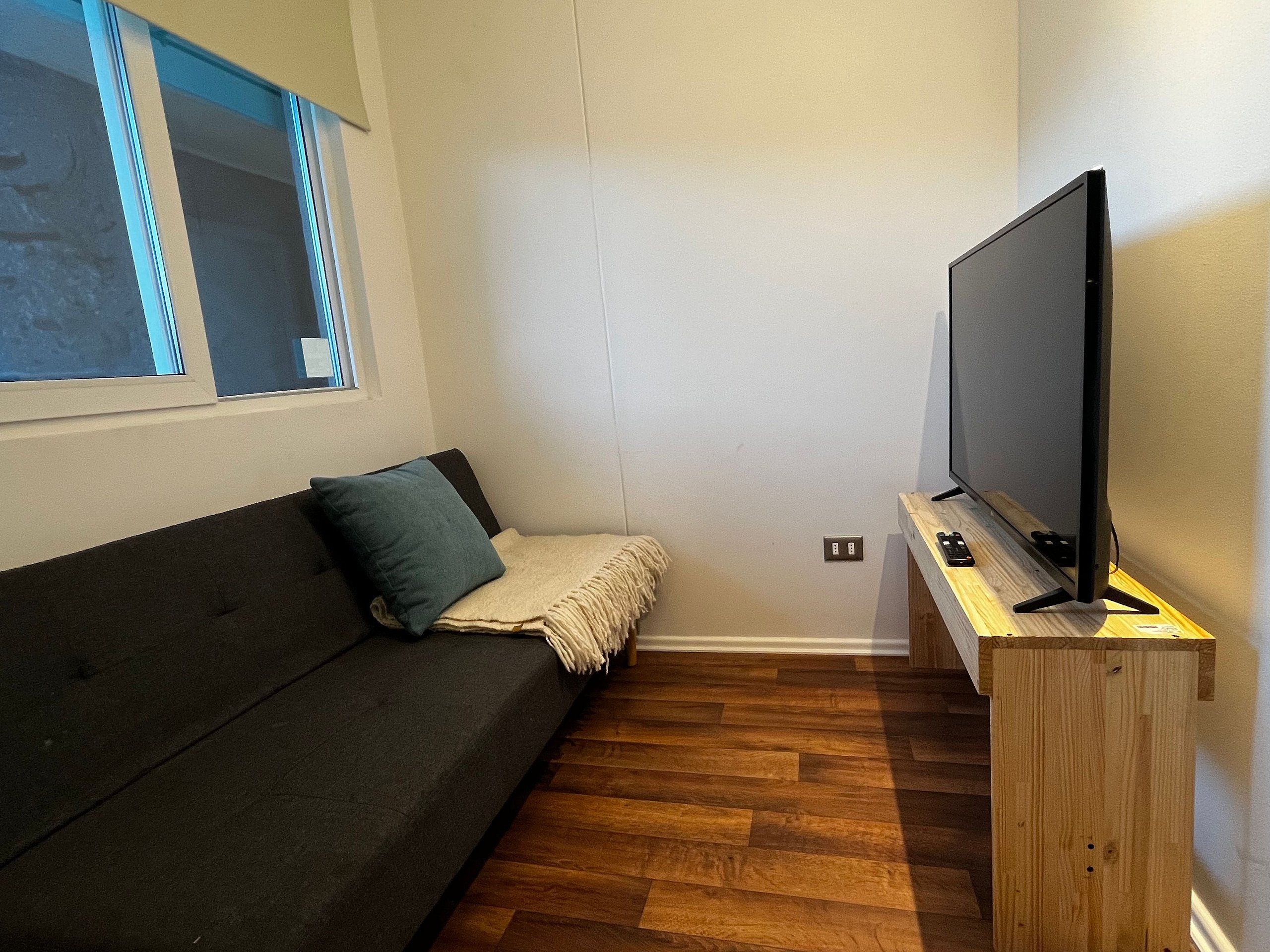 Ein kleiner Raum mit einem dunkelgrauen Sofa, einem Fernseher auf einem Holzschrank und einem Fenster mit Jalousien.