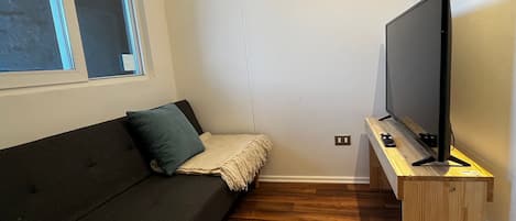A small room with a dark-colored sofa, a television on a wooden stand, and a window with blinds.