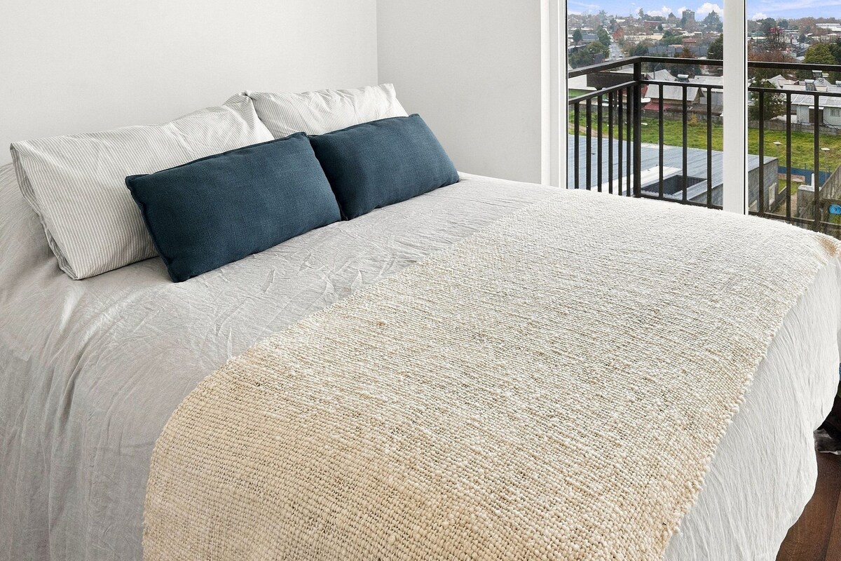 A bed with a white and beige blanket, two pillows, and a view of a cityscape through a balcony railing.
