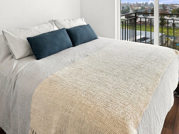 A bed with a white and beige blanket, two pillows, and a view of a cityscape through a balcony railing.