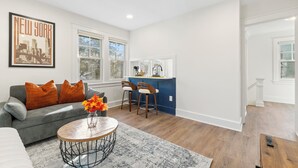 A spacious living room with a gray sofa, orange pillows, and a coffee table with a vase of flowers. There is a kitchen area with a blue accent wall and wooden bar stools. - The Bar Loft - Second Floor Suite. Close proximity to NYC and MetLife Stadium. (Bergenfield)