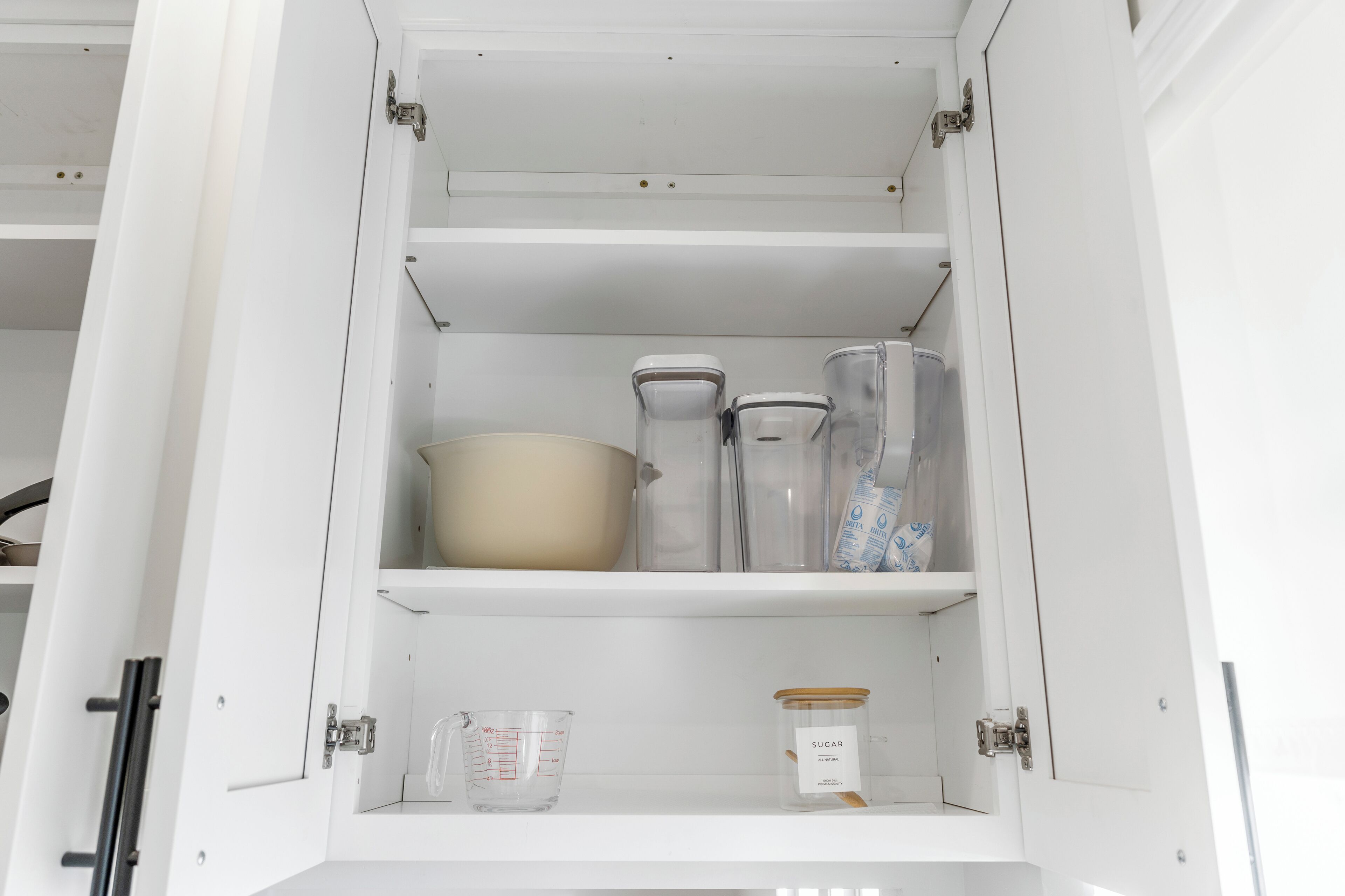 A kitchen cabinet with shelves containing a measuring cup, a bowl, and a jar of sugar.