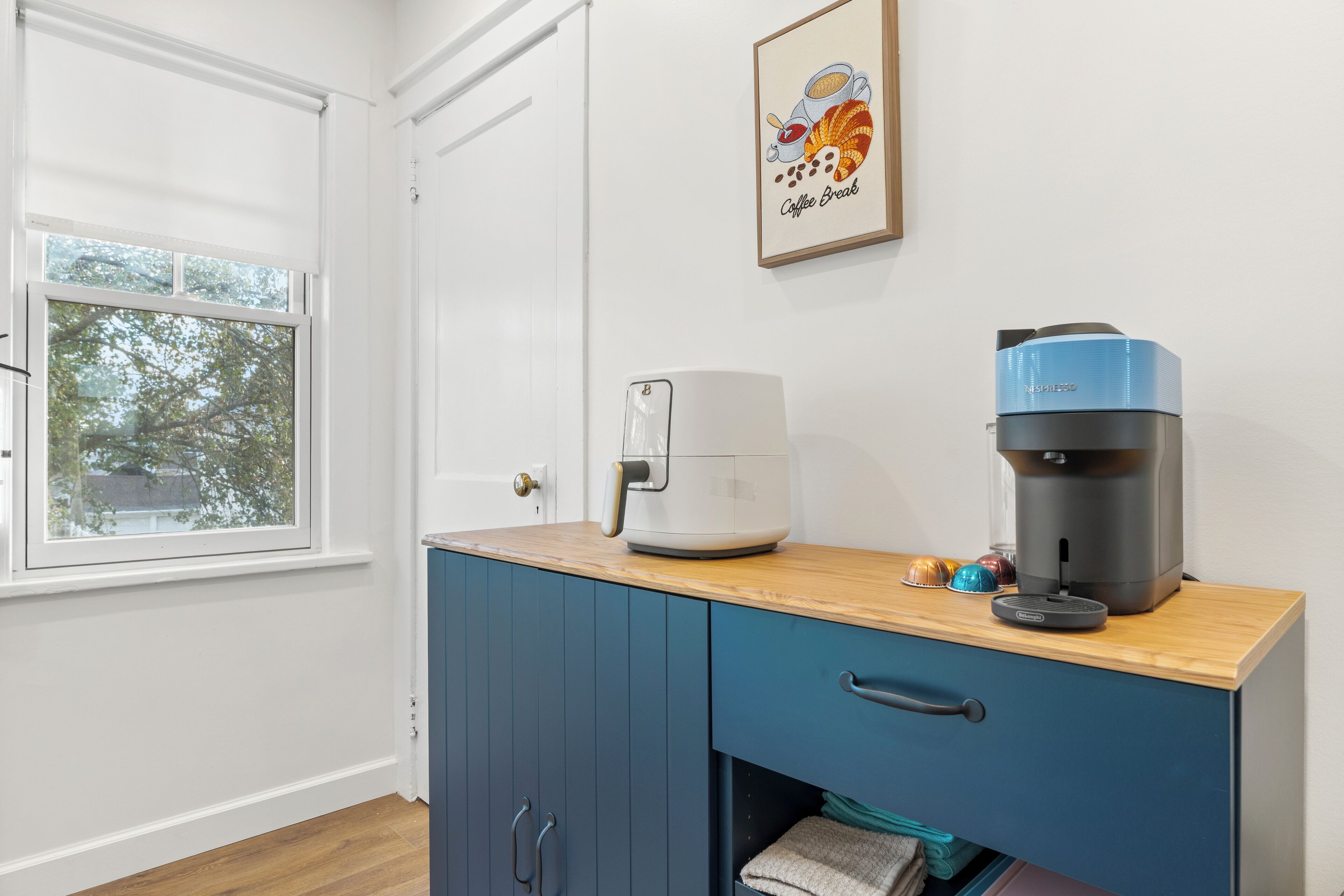 Une petite cuisine équipée d’une machine à café, d’une bouilloire et d’un tableau encadré au mur.