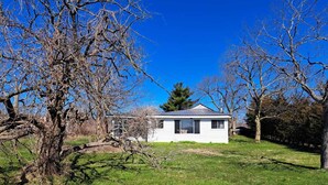 A small, single-story white house with a dark roof, surrounded by a grassy yard and leafless trees. - South Shore Cottage With Lake View (Pelee)