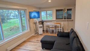 A modern living room with a large sectional sofa, a dining table with chairs, a flat-screen TV mounted on the wall, and a large window offering a view of the outdoors. - South Shore Cottage With Lake View (Pelee)