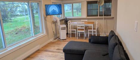 A modern living room with a large sectional sofa, a dining table with chairs, a flat-screen TV mounted on the wall, and a large window offering a view of the outdoors.