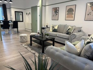 Un salon moderne avec un canapé gris, une table basse noire et un pouf blanc. On aperçoit à l’arrière-plan un coin repas avec une table et des chaises.