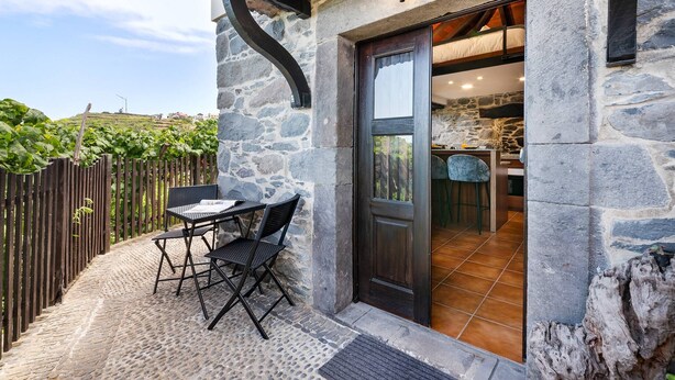 Un bâtiment en pierre avec une porte en bois sombre, un petit espace extérieur aménagé avec une table et des chaises, et une vue sur l’intérieur à travers la porte.