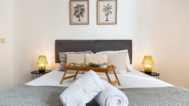 A neatly made bed with a grey blanket, two white towels, and a tray with toiletries. There are two wall-mounted lamps on either side of the bed.