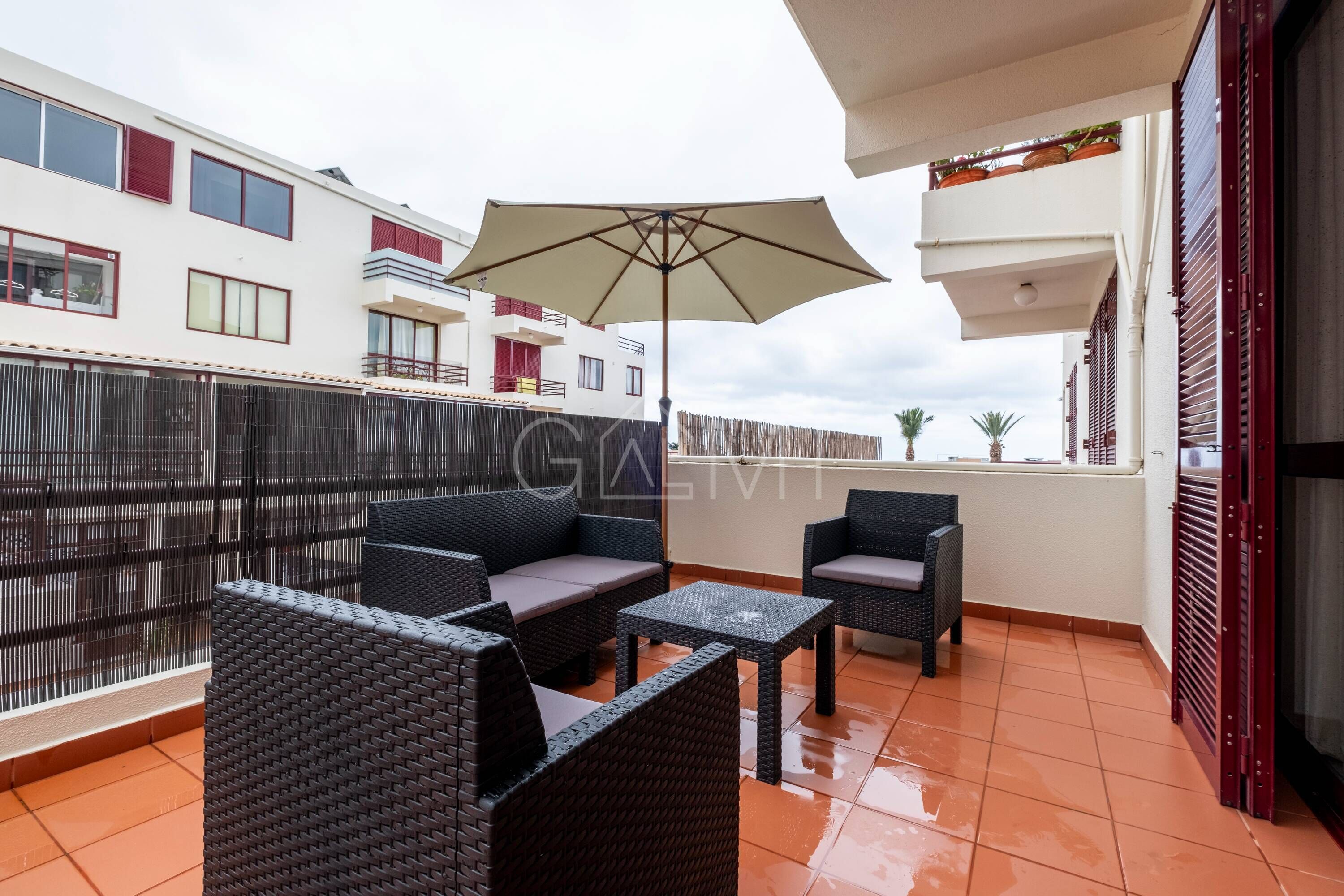 A balcony with wicker furniture, a large umbrella, and a view of neighboring buildings.
