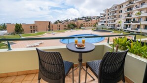 A balcony with two wicker chairs, a small round table with a plate of food and two glasses of orange juice, and a view of a swimming pool and buildings in the background. - Sarah's Apartment by GALMI (Santa Cruz)
