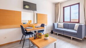 A modern living room with a wooden dining table, blue chairs, a sofa, a TV mounted on the wall, and a window with curtains. - Sarah's Apartment by GALMI (Santa Cruz)