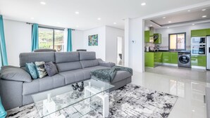 A modern living room with a grey sofa, glass coffee table, and a patterned rug. Adjacent is a kitchen with green cabinets and a washing machine. - Shell House by GALMI (Calheta (Madeira))