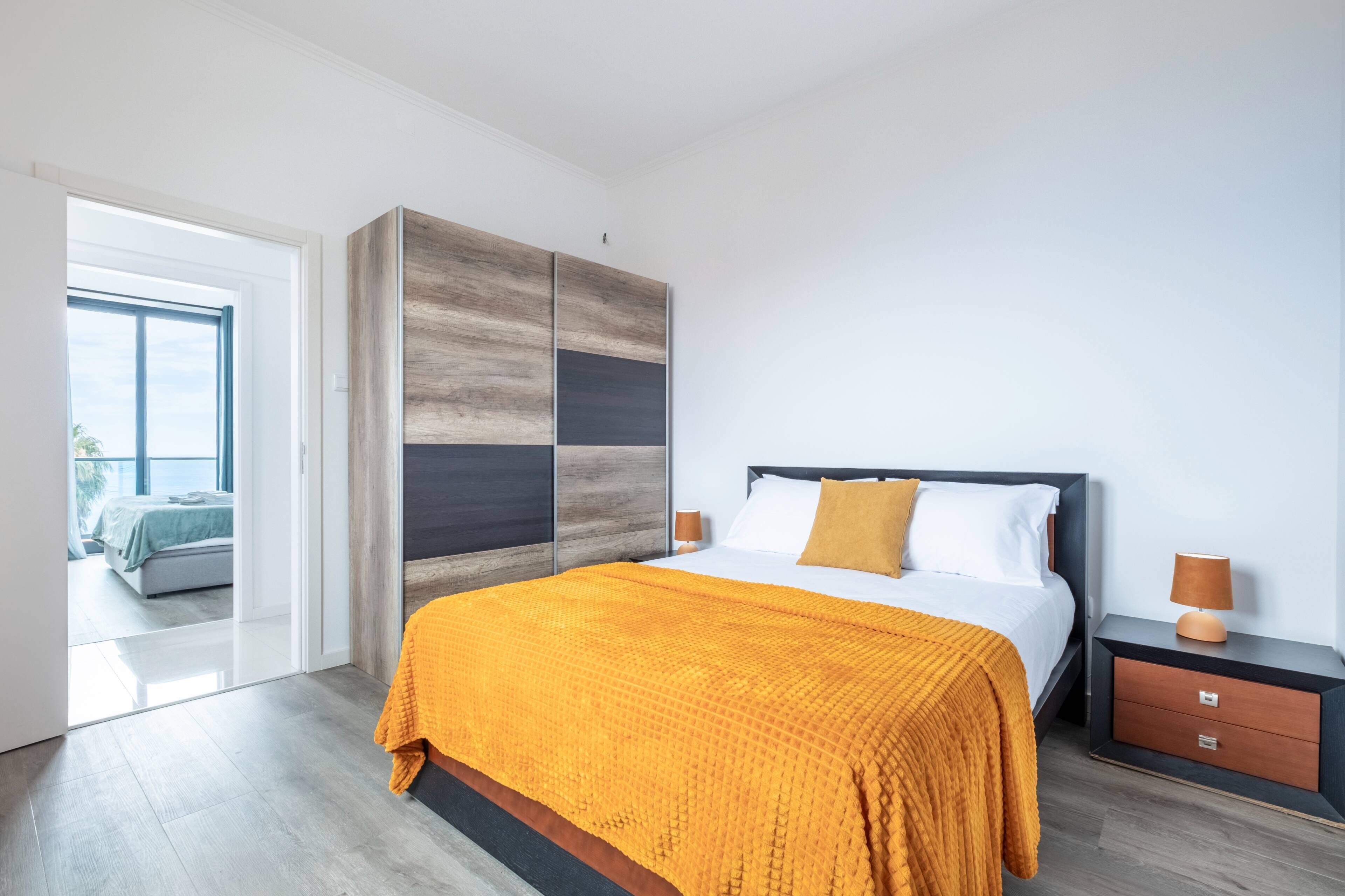 Une chambre moderne avec un grand lit, une armoire en bois, une table de chevet et une vue sur une autre pièce grâce à une porte ouverte.
