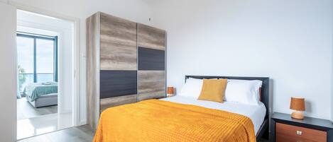 Une chambre moderne avec un grand lit, une armoire en bois, une table de chevet et une vue sur une autre pièce grâce à une porte ouverte.