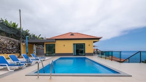 A swimming pool with blue lounge chairs, a building with a red-tiled roof, and a view of the sea. - Shell House by GALMI (Calheta (Madeira))
