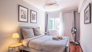 A neatly arranged bedroom with a bed, a bedside table with a lamp, a standing fan, and framed pictures on the wall. - Stylish Garden Apartment by GALMI (Santa Cruz)