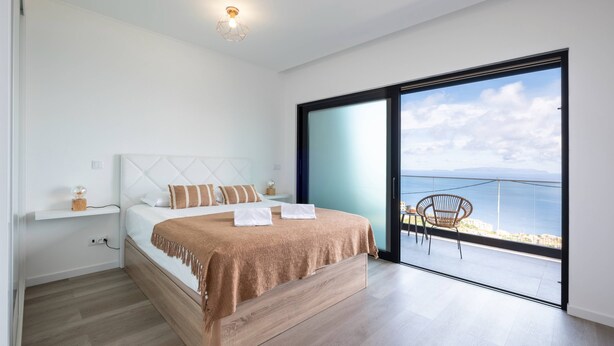 A modern bedroom with a large bed, a nightstand, a chair, and a view of the sea through a sliding glass door. - Villa Bela Horizonte by GALMI (Santa Cruz)