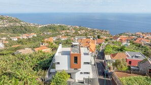 A coastal residential area with modern houses, a clear view of the sea, and a road leading to a building with a flat roof and a water tank.