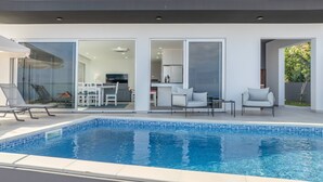 A modern outdoor pool area with a clear blue pool, a dining area with a table and chairs, and a kitchen visible through glass doors. - Villa Oceano Sereno by galmi (Ponta do Sol)