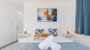 A neatly made bed with a blue blanket, two bedside tables with lamps, a tray with fruits and a bottle, and a painting on the wall. - Villa Oceano Sereno by galmi (Ponta do Sol)