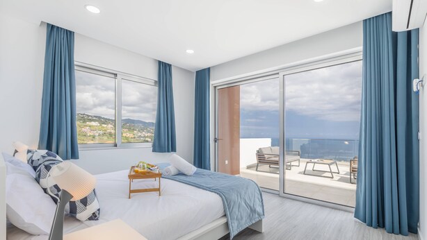 A modern bedroom with a large bed, a bedside table, a lamp, and a view of the city through large windows. - Villa Oceano Sereno by galmi (Ponta do Sol)