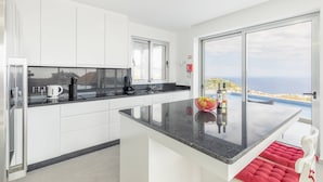 A modern kitchen with white cabinets, a black countertop, and a large window offering a view of the sea. - Villa Oceano Sereno by galmi (Ponta do Sol)