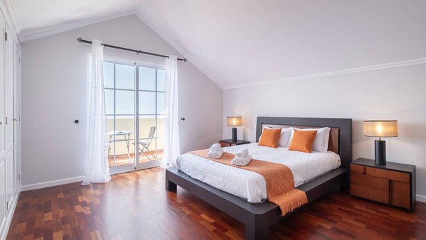 Ein modernes Schlafzimmer mit einem großen Bett, zwei Nachttischen mit Lampen, einem Balkon mit einem Sessel und einem Holzboden.