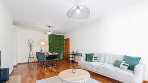 Una sala de estar moderna con un sofá blanco, una mesa de centro redonda de madera y una zona de comedor con una pared verde y suelo de madera.