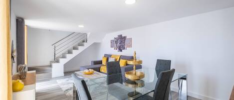 A modern dining area with a glass table and chairs, a staircase leading to another level, and a Buddha statue on the wall.