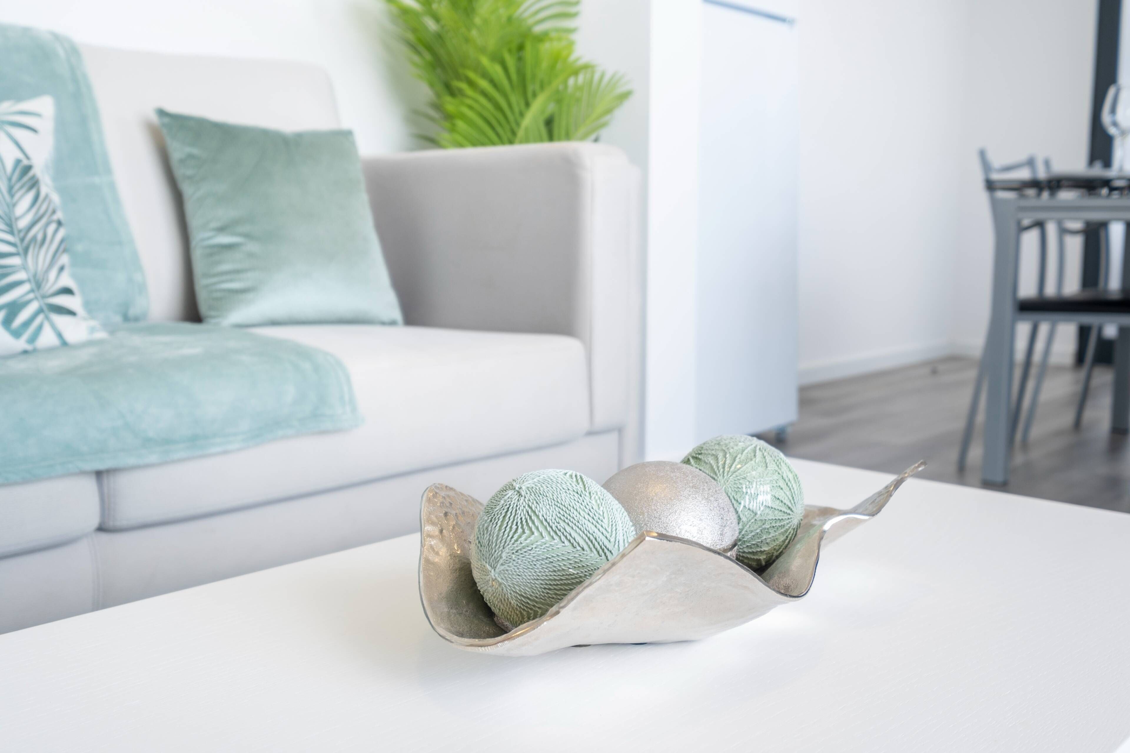 A modern living room with a white sofa, a decorative metal bowl with yarn balls, and a dining area in the background.