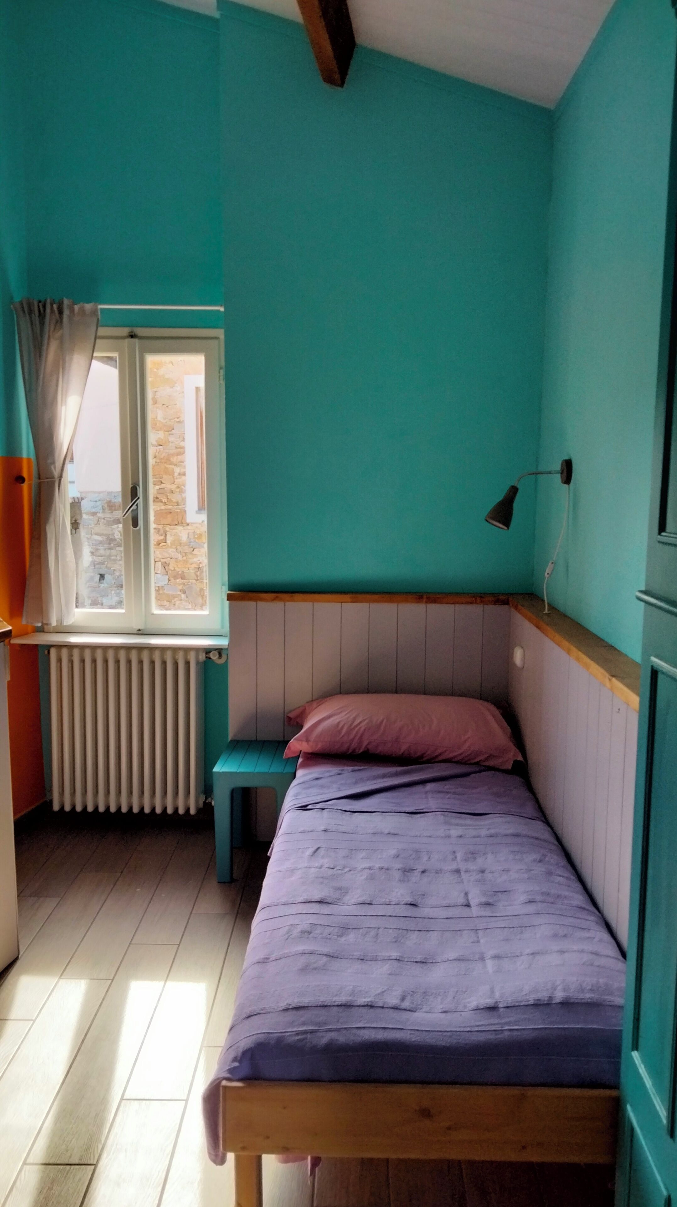 Een kleine kamer met een enkel bed, teal muren, een houten hoofdbord, een raam met gordijnen, een radiator en een bedlampje.