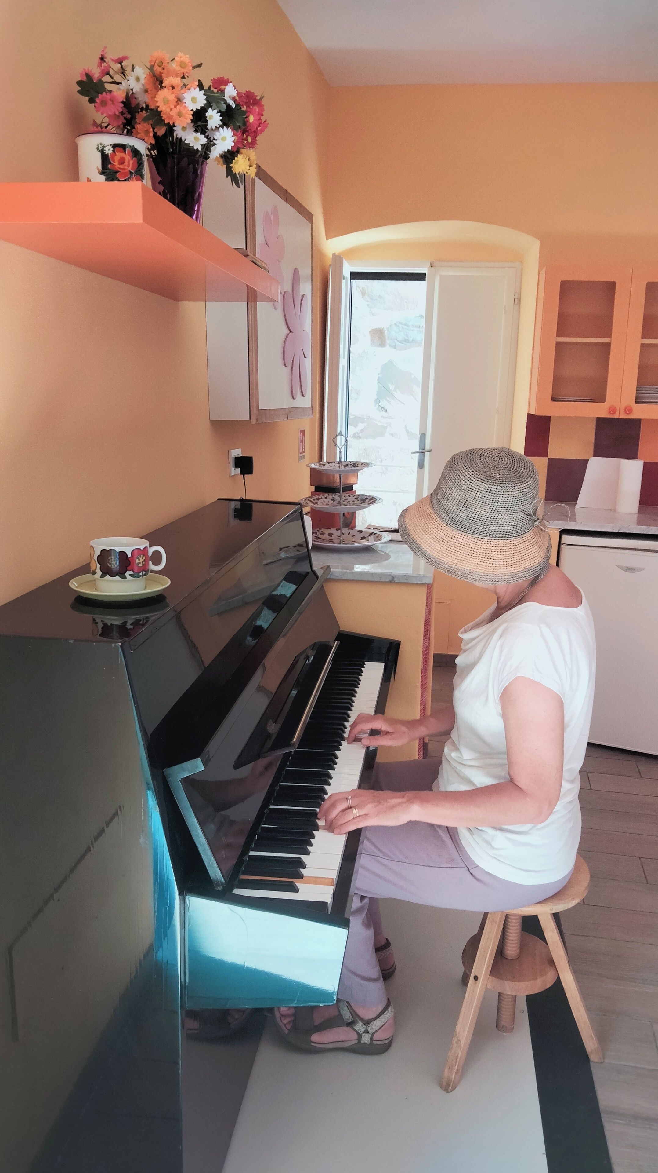 Eine Person spielt Klavier in einem Zimmer mit Fenster, kleinem Kühlschrank und einer Dekovase mit Blumen auf einem Regal.