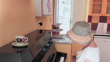 Eine Person spielt Klavier in einem Zimmer mit Fenster, kleinem Kühlschrank und einer Dekovase mit Blumen auf einem Regal.