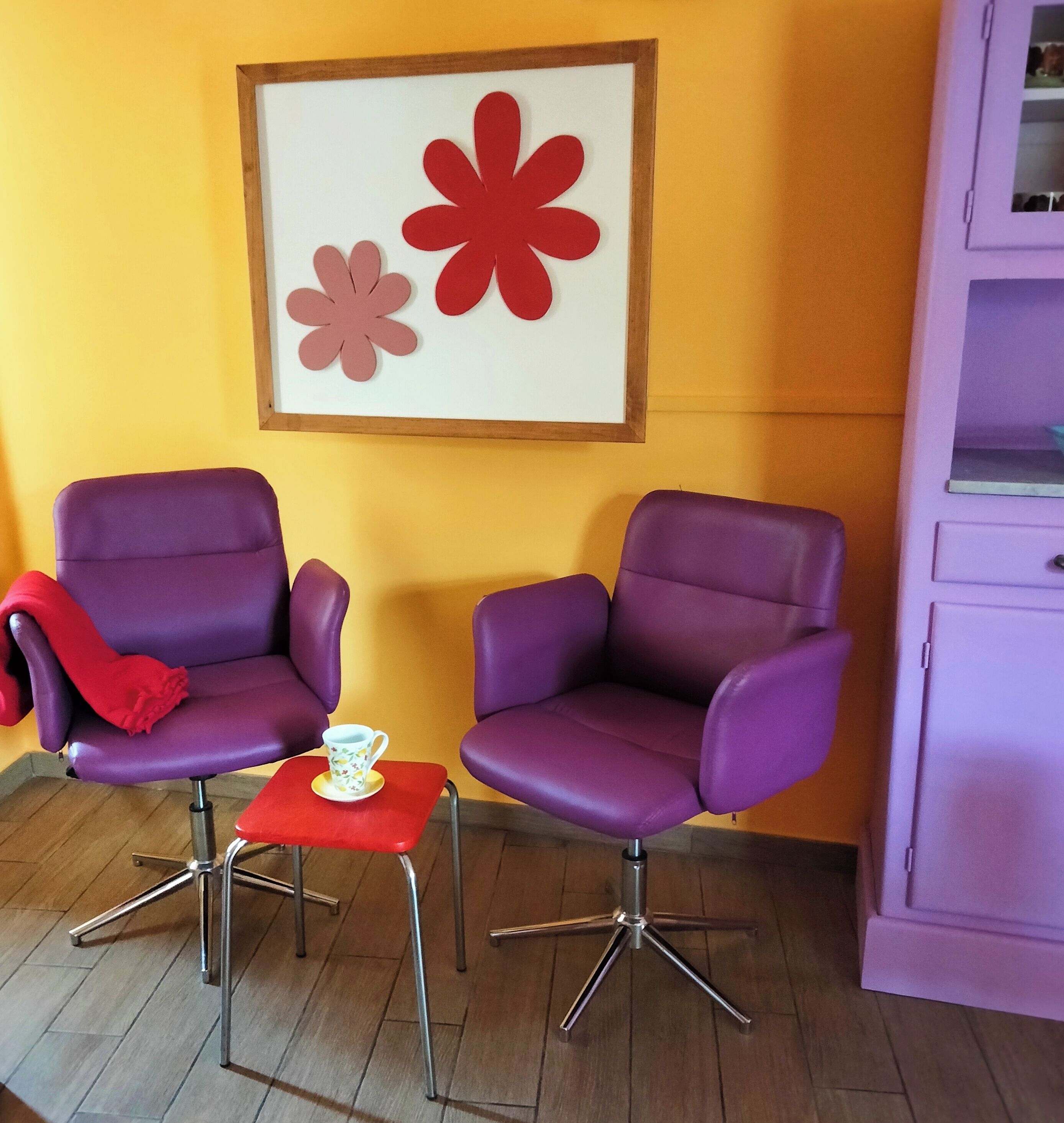 Two purple armchairs, a small red table with a white cup, and a framed artwork with red flowers on a yellow wall.