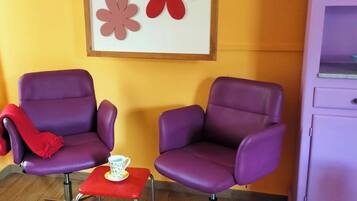 Two purple armchairs, a small red table with a white cup, and a framed artwork with red flowers on a yellow wall.