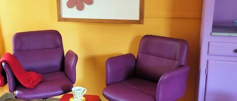 Two purple armchairs, a small red table with a white cup, and a framed artwork with red flowers on a yellow wall.