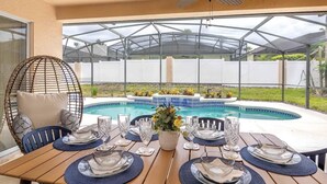 Un área de comedor al aire libre techada, con piscina, ventilador de techo y una silla con cojines.