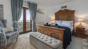A bedroom with a bed, a wooden dresser, a bench, a chair, and a balcony with curtains.