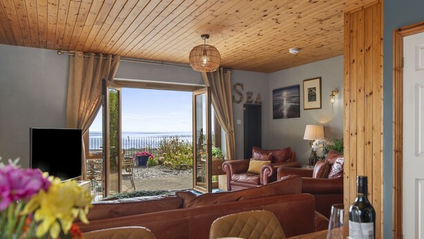 A room with a wooden ceiling, a sofa, a dining table with a bottle and glasses, and a view of the sea.