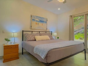 A bedroom with a bed, a nightstand, a dresser, a ceiling fan, and a window with a view of greenery.