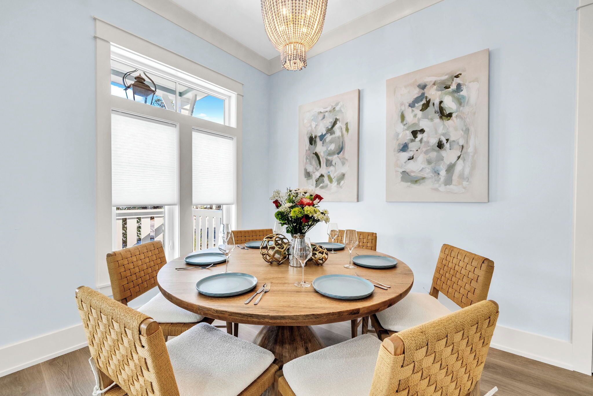 A dining area with a round wooden table, six woven chairs, and a vase of flowers. There are abstract paintings on the walls and a chandelier above.