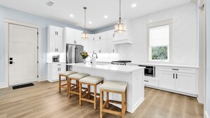 Une cuisine moderne avec des meubles blancs, un îlot central doté de tabourets, des appareils en acier inoxydable et un éclairage suspendu.